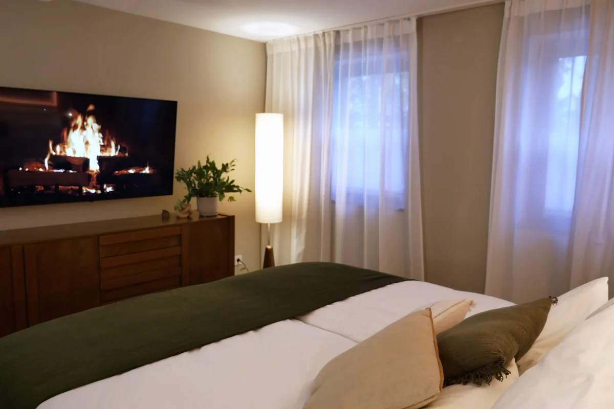 Schlafzimmer der WolkenGuckerei am Abend, im Fernseher an der Wand brennt ein Kaminfeuer, davor das Bett mit grüner Tagesdecke