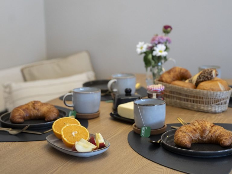 Esstisch Tisch gedeckt mit Croissants, Orangenscheiben, Tassen und einer Blumenvase.