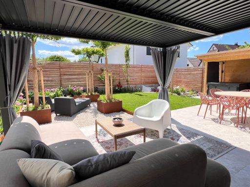 Grüner Garten mit Sitzbereich, Terrassenschirm und blühenden Pflanzen.
