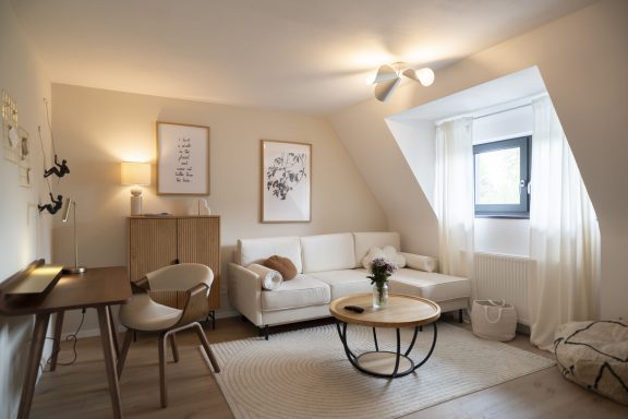 Wohnzimmer WolkenBlick Heller Wohnraum mit beigem Sofa, Holztisch und Fenster, dekoriert mit Pflanzen und Kunst.