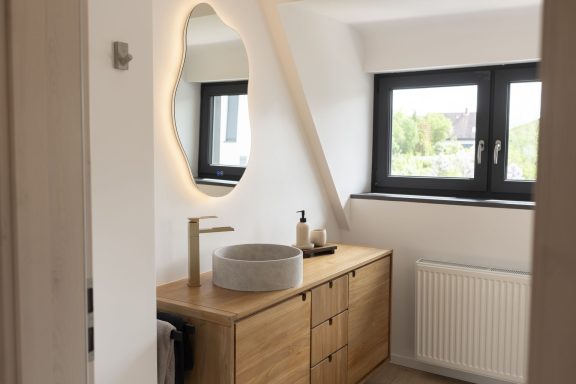 Bad WolkenBlick Modernes Badezimmer mit Holzschrank, rundem Waschbecken und beleuchtetem Spiegel.