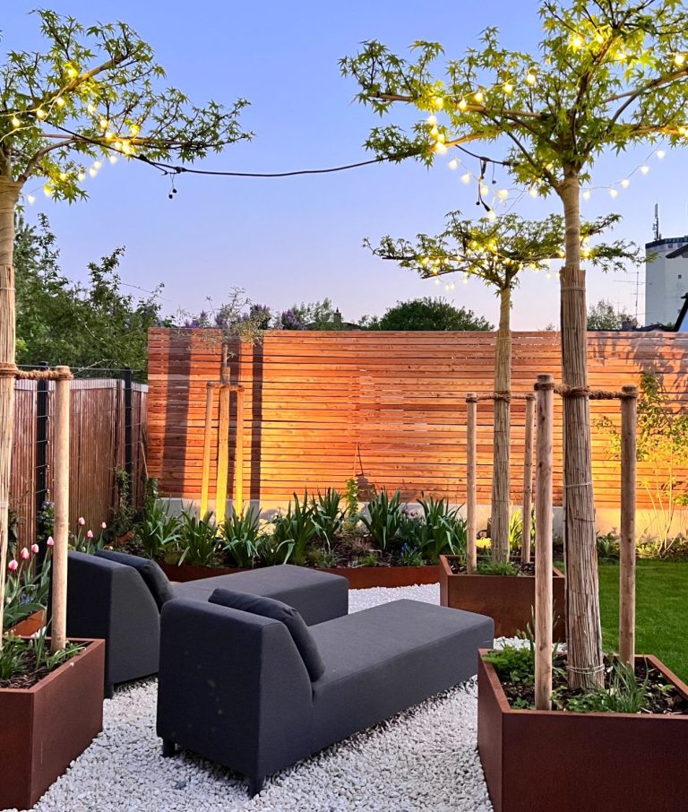 Moderne Sitzecke im Garten mit Holzzaun, Bäumen und Lichtern.