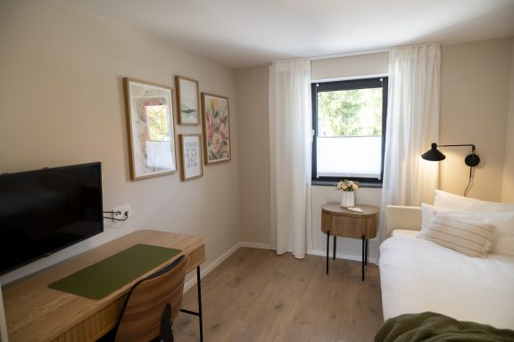 Schnelles WLAN Helles Zimmer mit Bett, Schreibtisch, Stuhl und Fernseher vor einem Fenster.