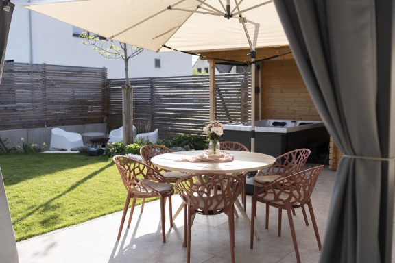 Gemütlicher Außenbereich mit Tisch, Stühlen und Sonnenschirm, umgeben von Rasen und Holzlattenzaun.