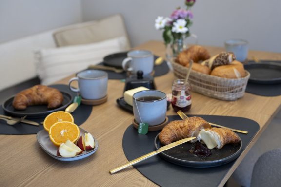 Geschirr Frühstückstisch mit Croissants, Orangenscheiben, Tassen und Blumen in der Mitte.