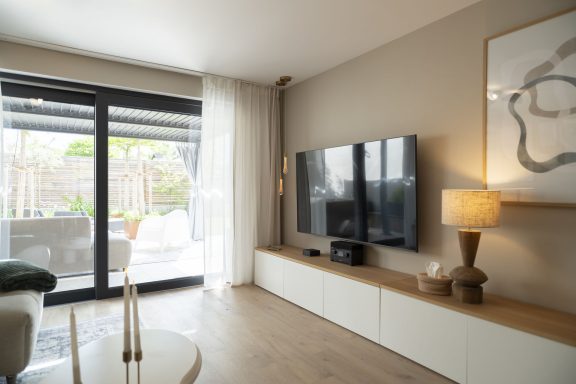 SKY Modernes Wohnzimmer mit Fernseher, Lampe und großen Fenstern, die auf einen Garten blicken.