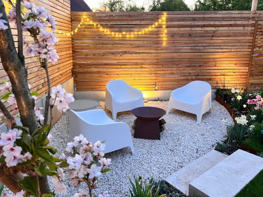 Gemütliche Gartenlounge mit weißen Stühlen, Tisch und blühenden Pflanzen.