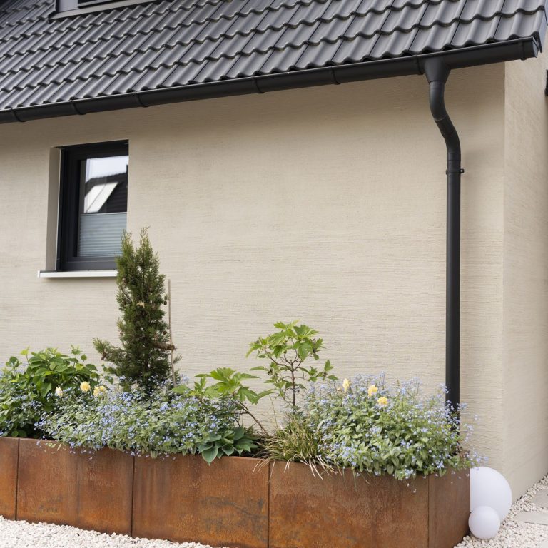 Hauswand mit bepflanztem Hochbeet, Blumen und kleinen Bäumen.