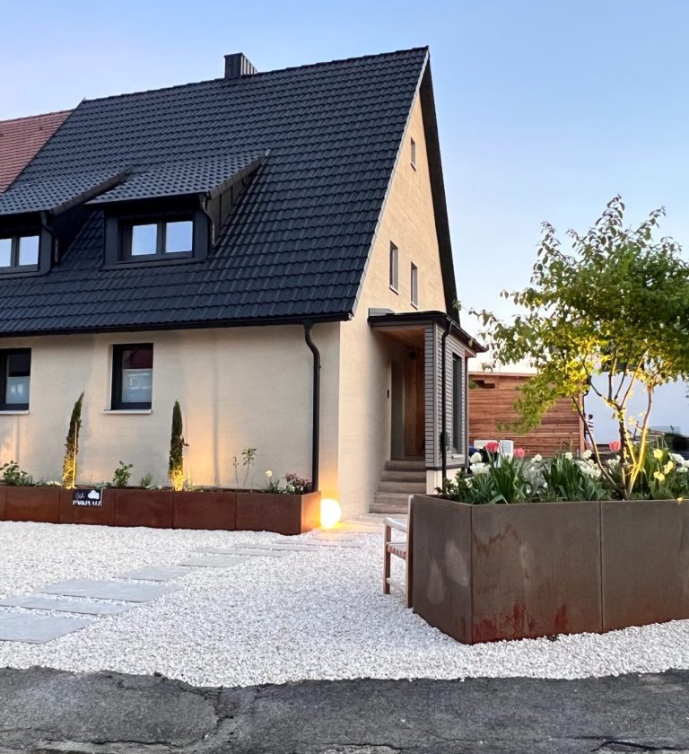 Helles Einfamilienhaus mit schwarzem Dach und schlichtem Außenbereich, umgeben von Bäumen.