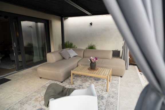 Überdachter Loungebereich Gemütliche Terrasse mit einer beigen Couch, Holztisch und Dekoration.
