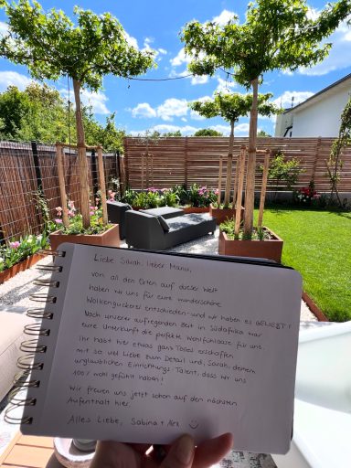 Handschriftliche Notizen vor einem gepflegten Garten mit Bäumen und Sitzbereich.